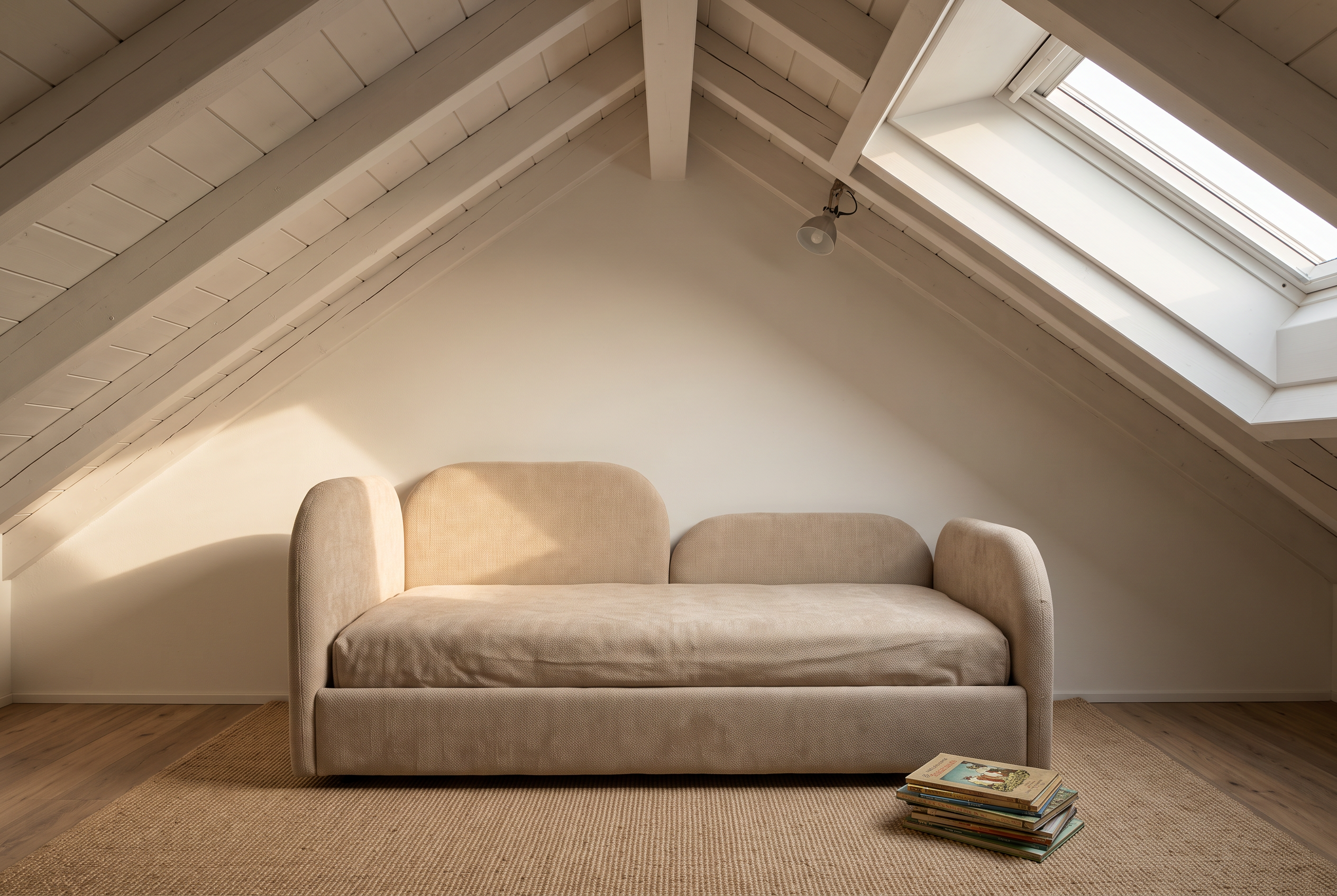 Niedriges gepolstertes Kinderbett in Sandbeige unter der Dachschräge eines Estrichzimmers mit Dachflächenfenster und Sisalteppich